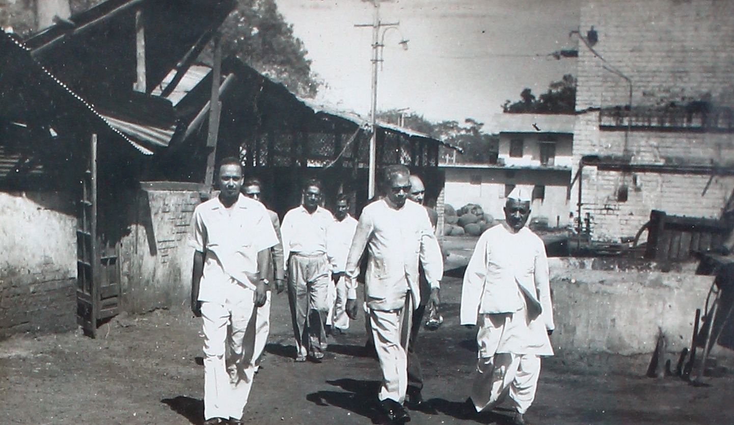 A group walking through the mill yard, sheds and a chimney behind them.