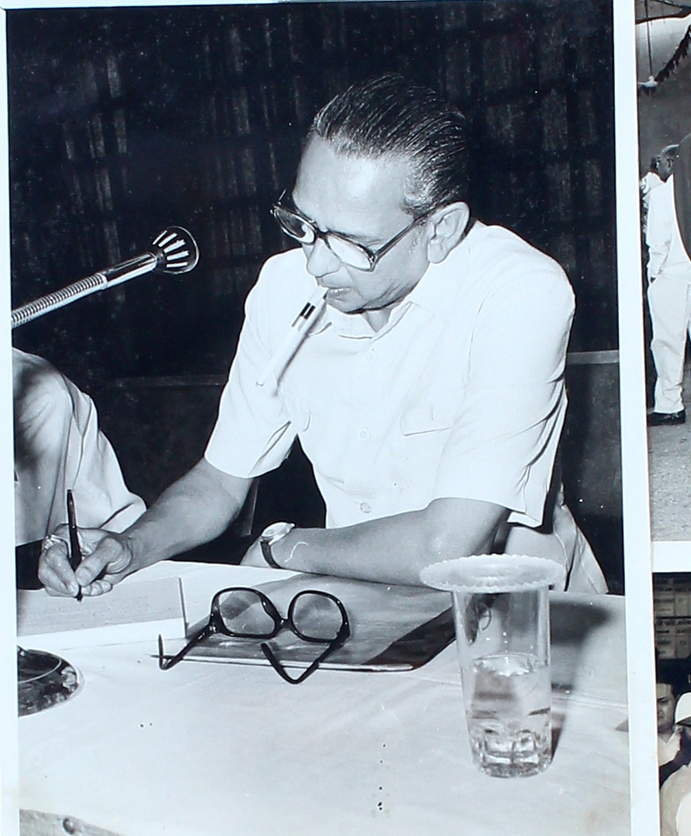 A man signing papers at a desk, spectacles and a glass of water beside him.